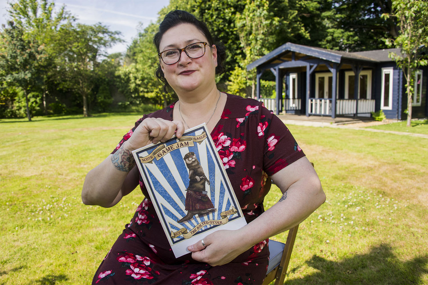 The image shows Charly Harbord holding a Kilted Otter poster whilst sitting in a garden.