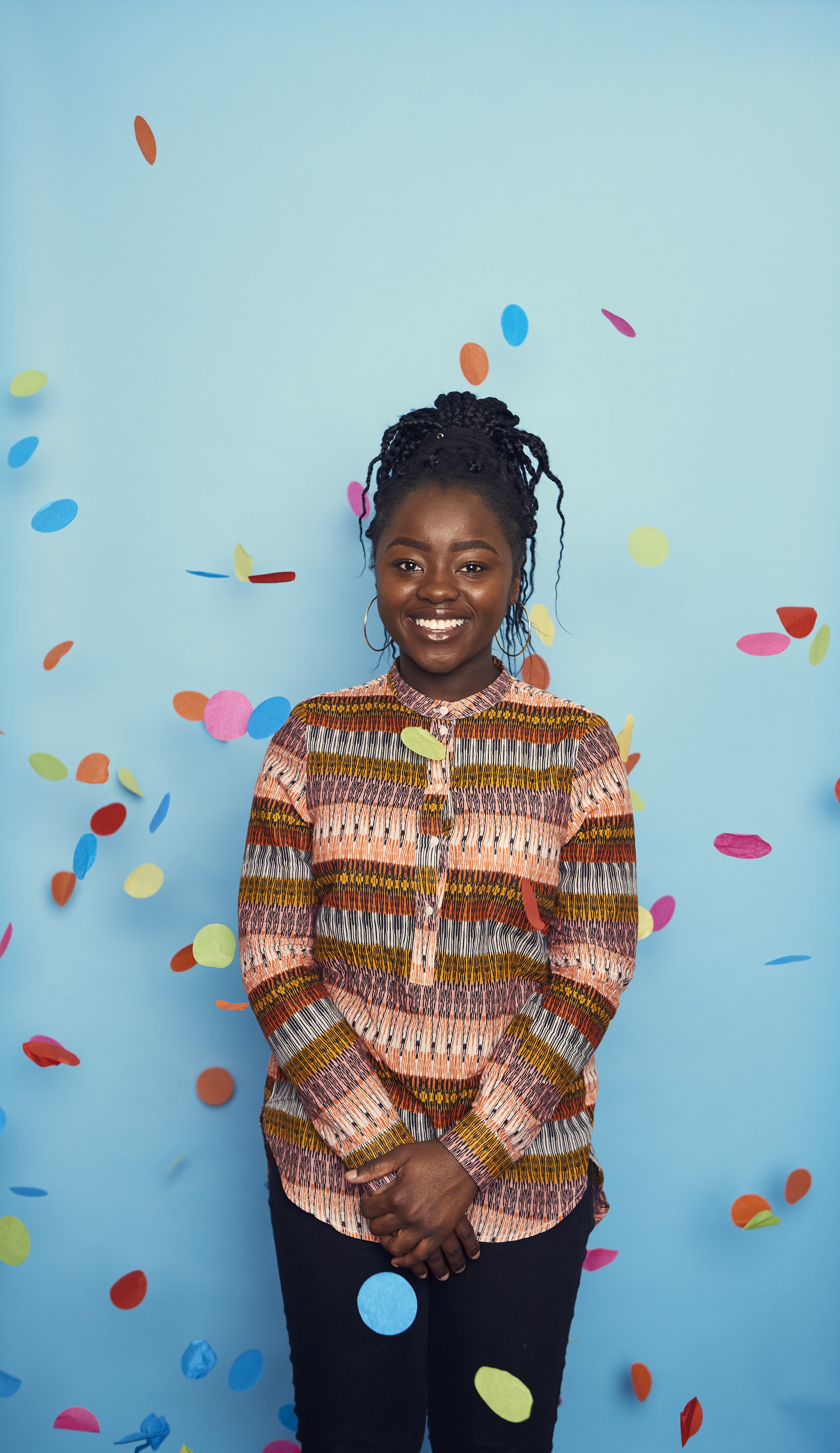 Smiling female standing in front of a blue background - lots of colorful bits of circle shaped paper are flying about in the air around her
