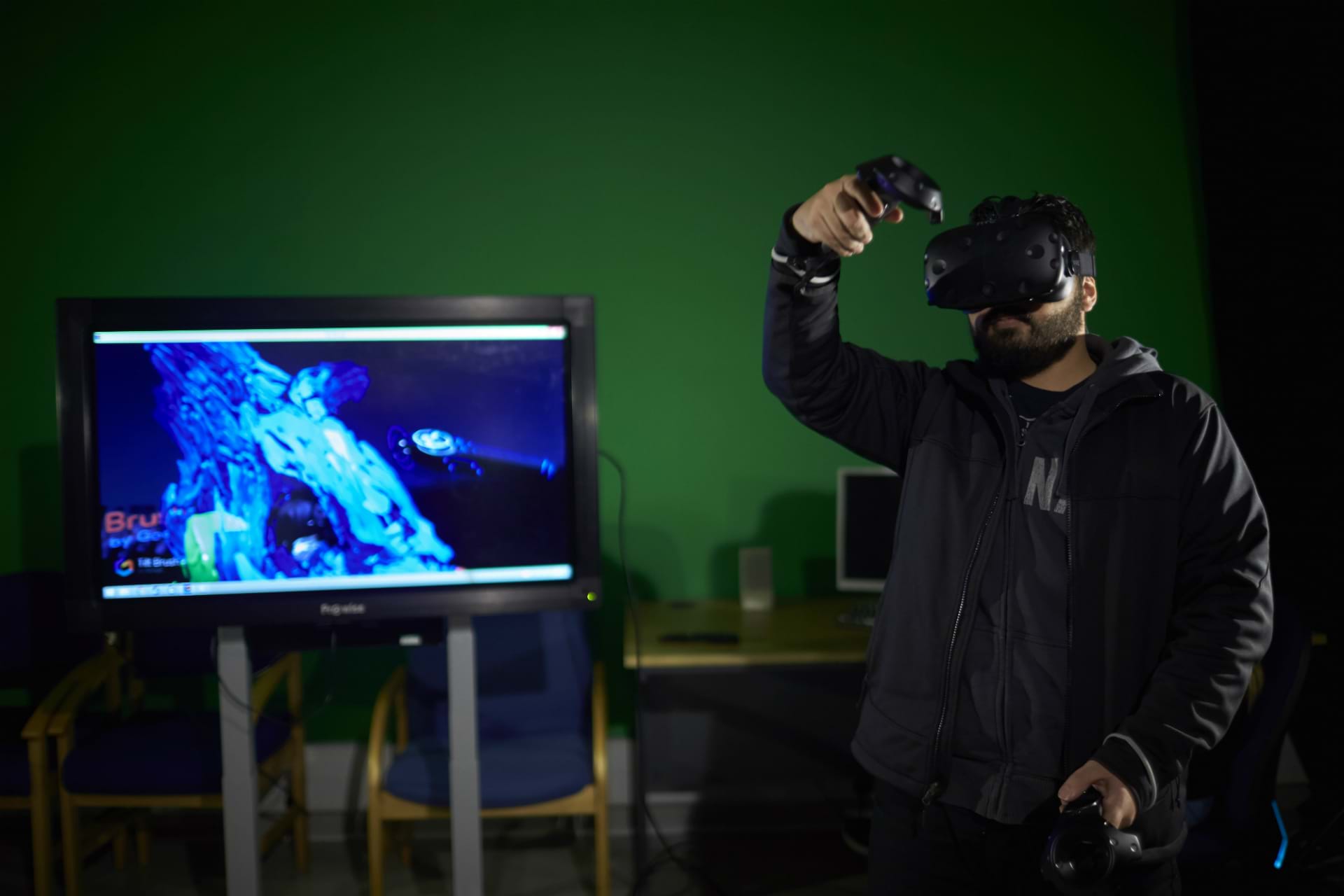 A student using VR in a green screen room with a screen in the background showing a video game
