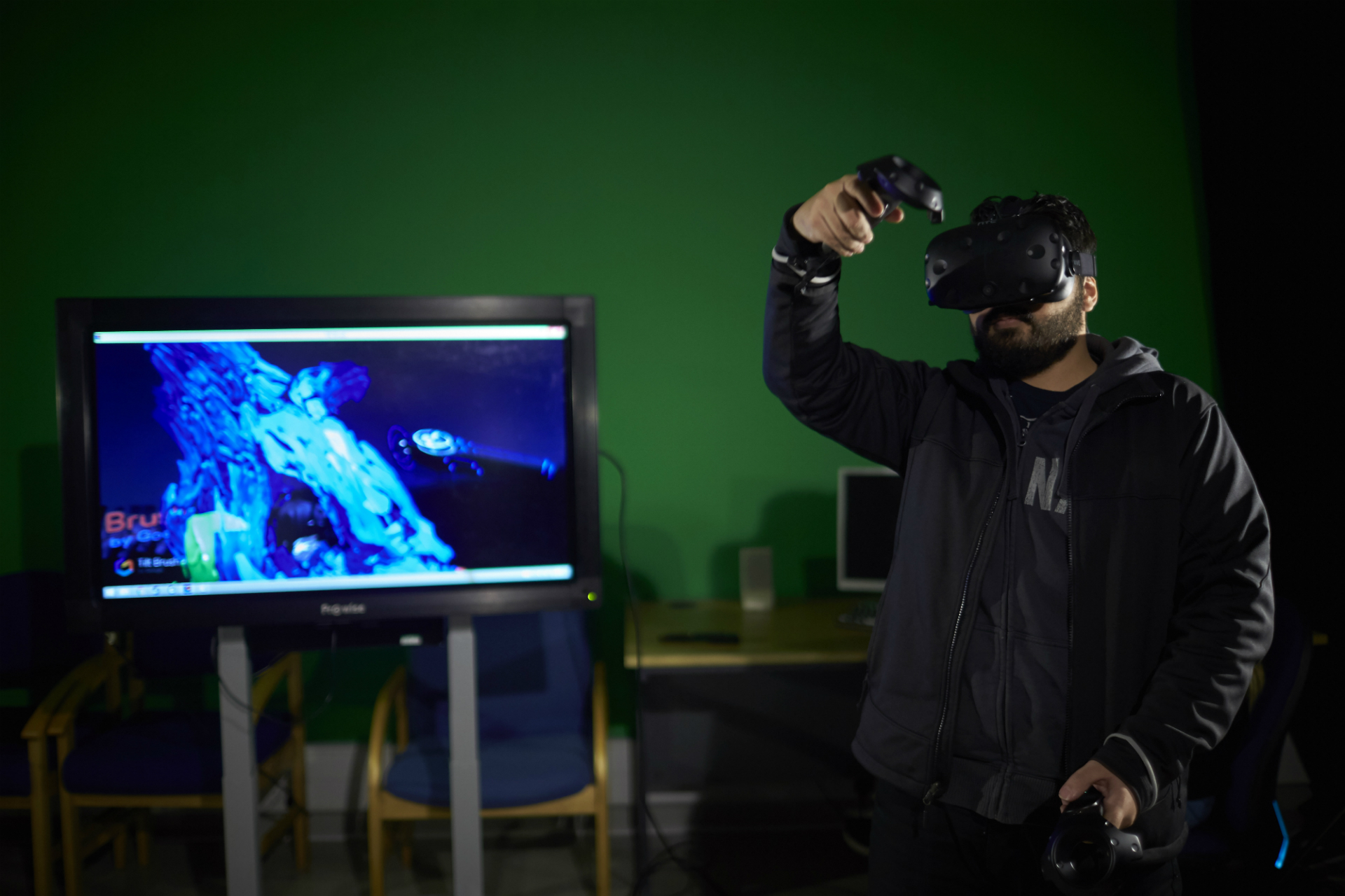 A student using VR in a green screen room with a screen in the background showing a video game