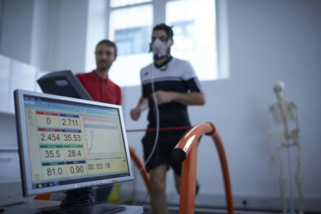 Male wearing breathing apparatus whilst running on a running machine - another male is standing beside him watching