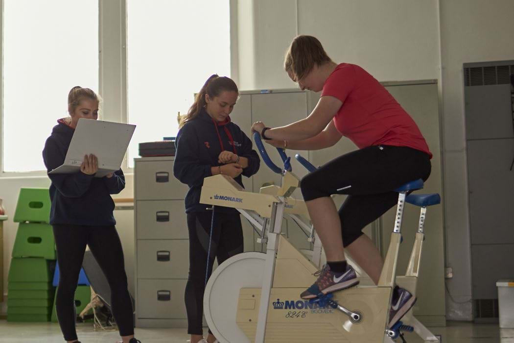 Female working on an exercise bike - two other females spectating and taking notes