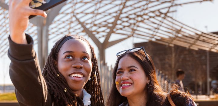 Two students outside standing next to each other looking up at a phone taking a selfie together. 