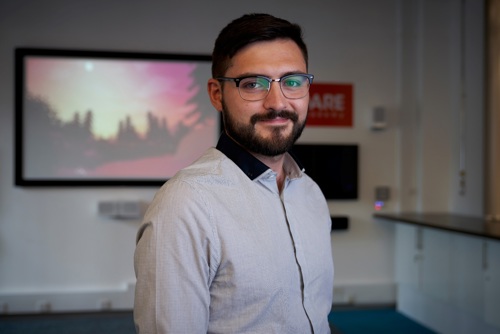 The image is a head shot of Alex Tarvert who is wearing a dress shirt and glasses. Behind him there is an image of a forest on a projector screen.