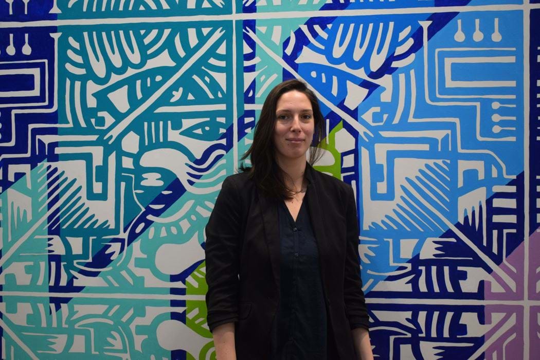 Picture of Sarah Quenel smiling, she is standing in front of a wall painted with a colourful pattern