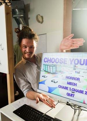 Picture of Cameron Moody presenting a project, gesturing towards a monitor with the words "Choose your game: Horror, Mech, Chase 1, Chase 2, Quit" on it.