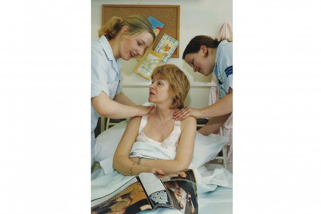 Two female nurses from Dundee College of Technology (Abertay university) on either side of a female patient. Nurses are propping the patient up with a pillow. 