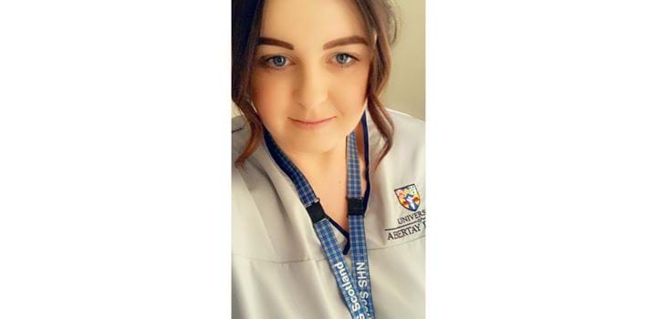 A selfie of Caroline Ford, in a nurses uniform with an Abertay University logo on her right. Caroline is also wearing an NHS Scotland lanyard arount their neck.