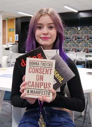 The image shows Student President Olivia Robertson in the Library holding up a range of books on Gender-Based Violence. - the book in the foreground is titled Consent on Campus.