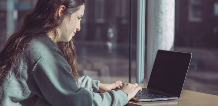 A woman with dark hair wearing a green sweatshirt, she sits at a desk working on a laptop with a window in front of her. 