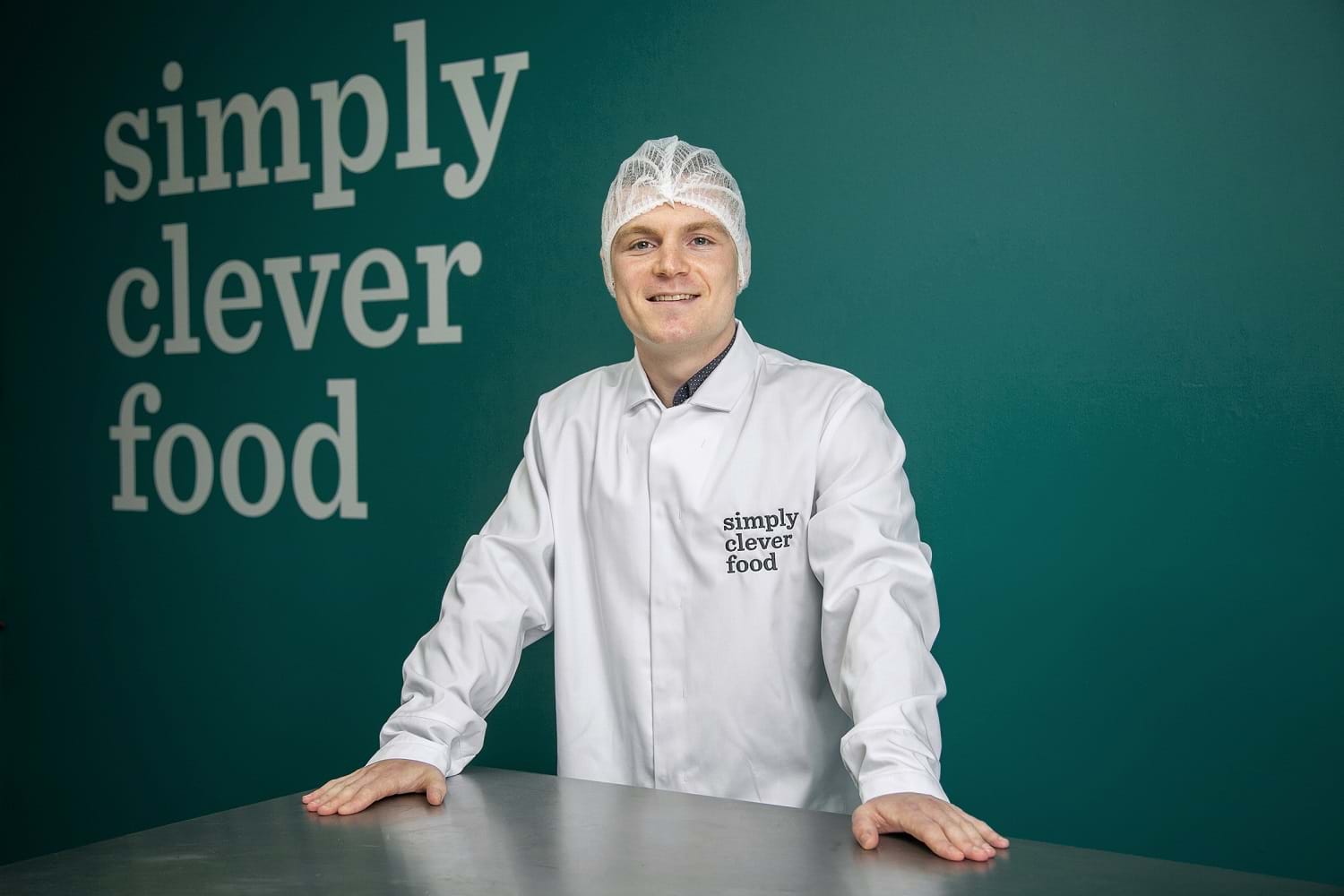 The image features Jamie in a lab coat and hairnet in front of a wall which has text reading "simply clever food".