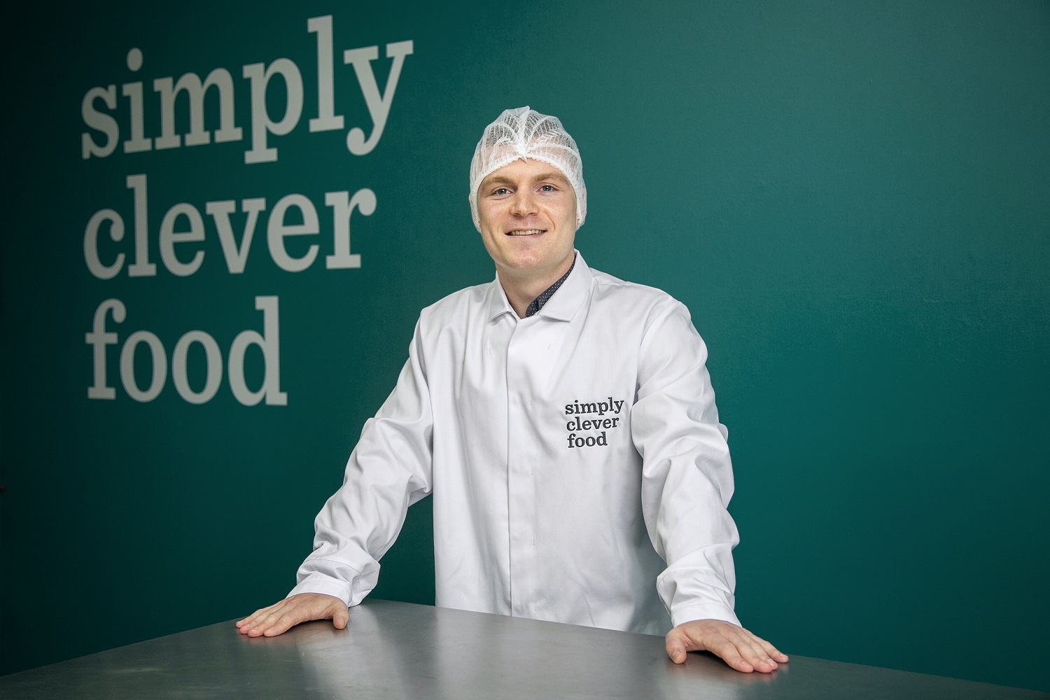 The image features Jamie in a lab coat and hairnet in front of a wall which has text reading "simply clever food".