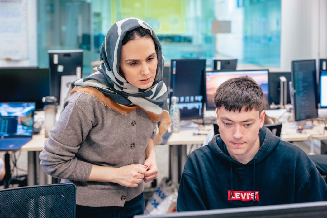 Two people looking at a computer screen in a classroom setting, there are computers seen in the background. 