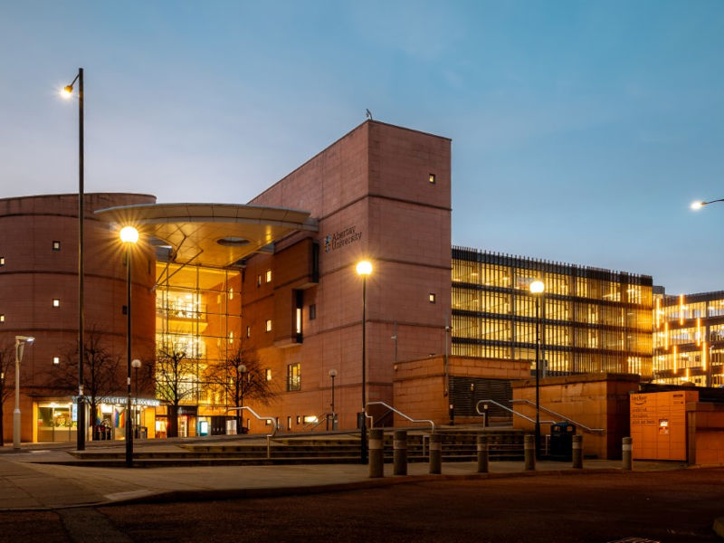 The Bernard King Library at Abertay University in Dundee, Scotland. The award-winning building features a distinctive curved glass façade resembling an open book, supported by a rectangular sandstone spine. The four-storey modern structure combines steel, glass, and stone elements in its contemporary architectural design.