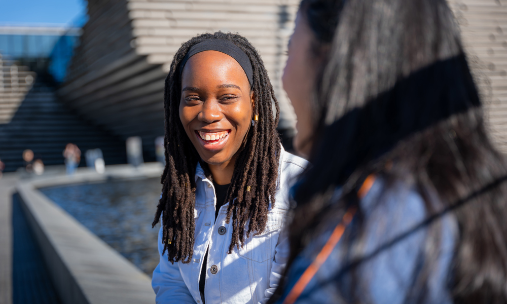 A woman sitting outside laughing and chatting to another woman sitting in front of her. 