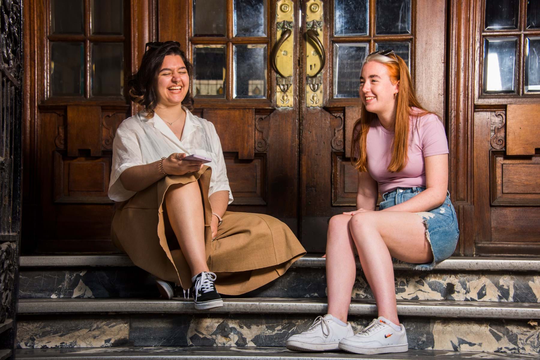 The image shows two students in summer clothes sitting on some marble steps in front of some wooden doors.